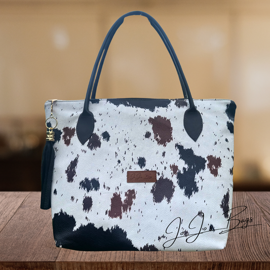 A cow-print tote bag with a white base, black and brown spots, dark navy handles, and a small brown leather logo patch, displayed on a wooden surface with a blurred warm background.