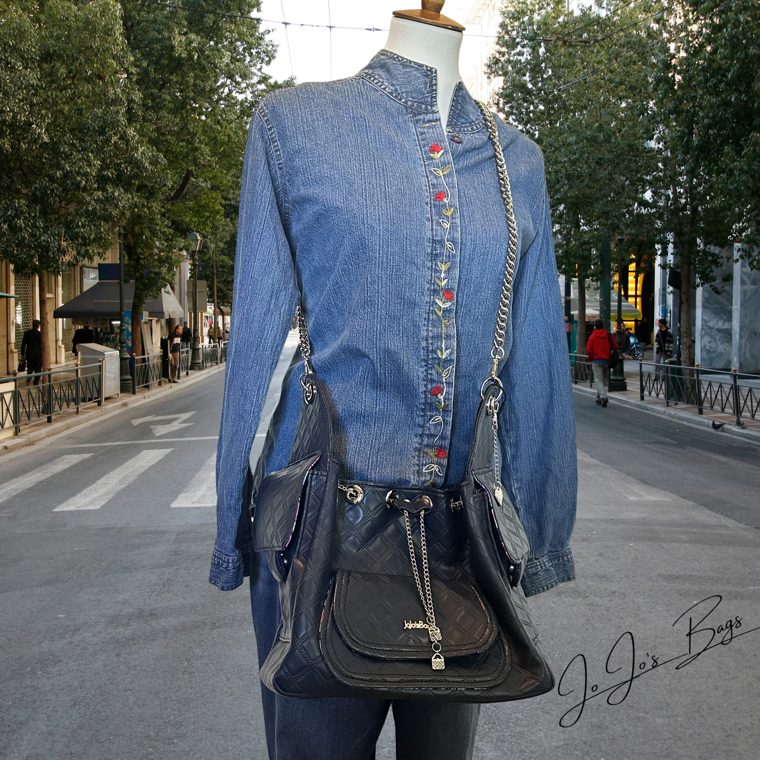 A mannequin dressed in a blue denim shirt and jeans wears a dark blue faux leather shoulder bag with silver chain straps, photographed on a city street with trees and buildings in the background.