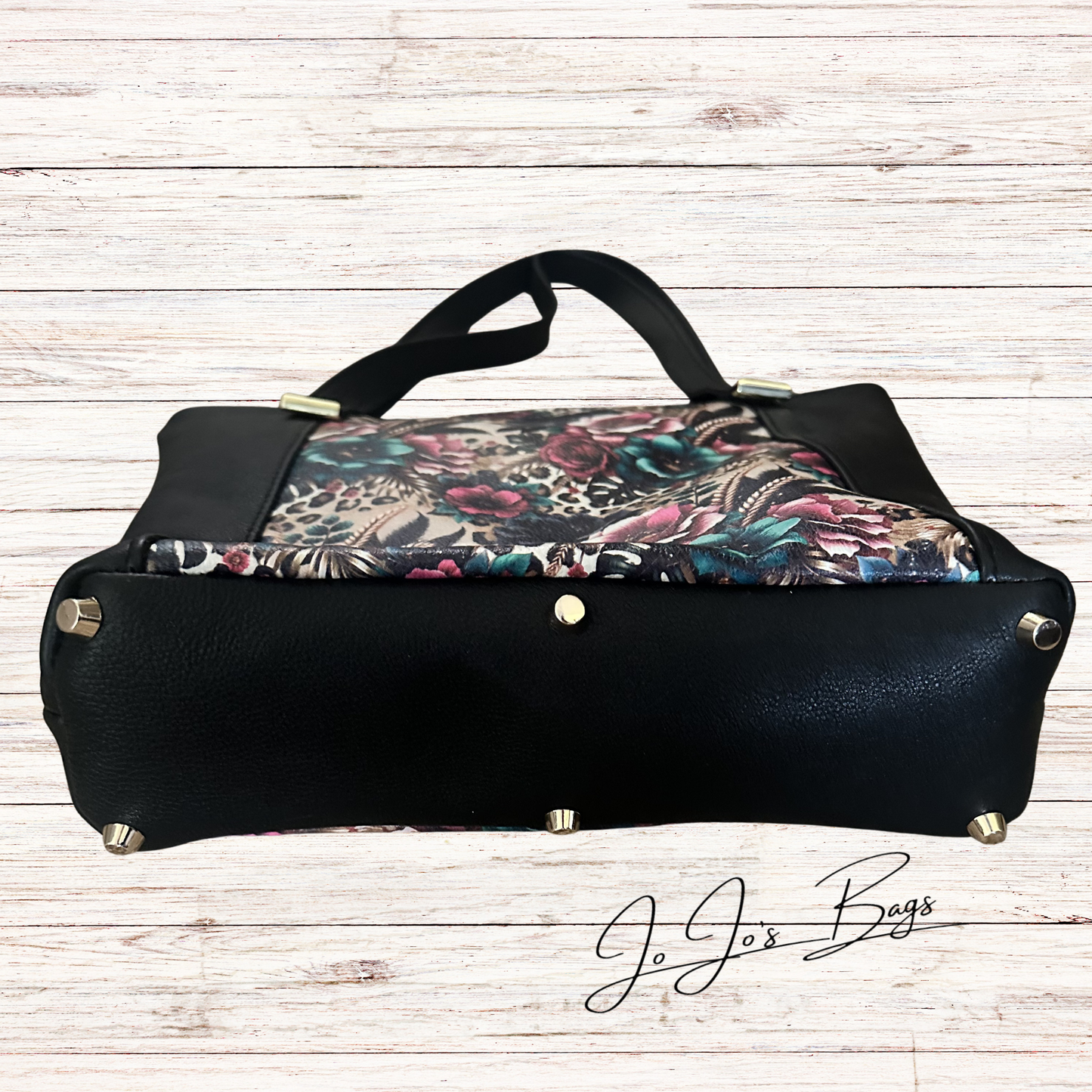 A black leather handbag with a floral-patterned fabric insert, displayed on a light wood plank background. The bag features a black handle, gold-tone studs, and a signature watermark for 'Jo Jo's Bags' in the lower right corner.