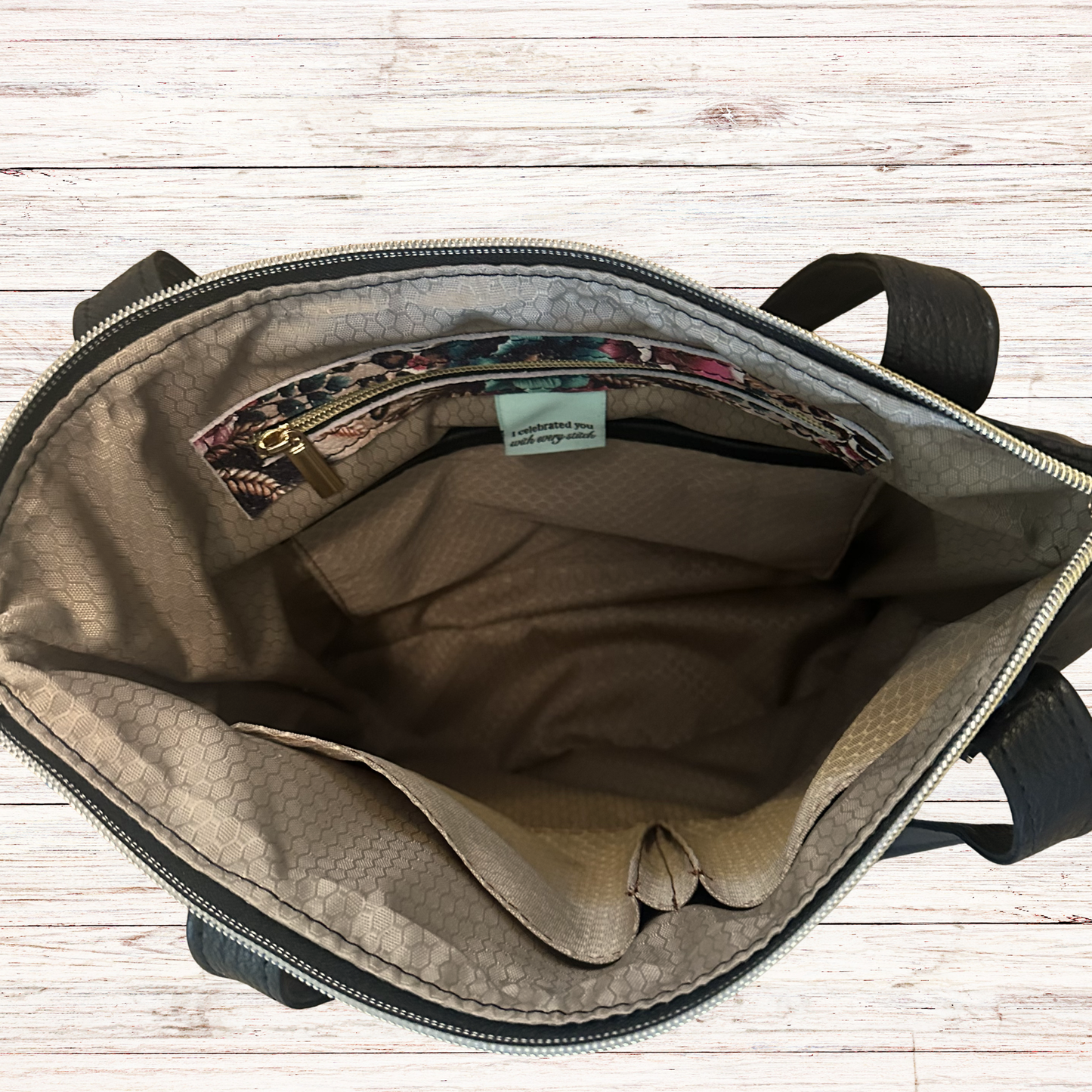 An open bag shown from above, revealing a beige and brown patterned interior with a zippered pocket and a small light blue label. The bag has black handles and is photographed on a light wood plank surface.