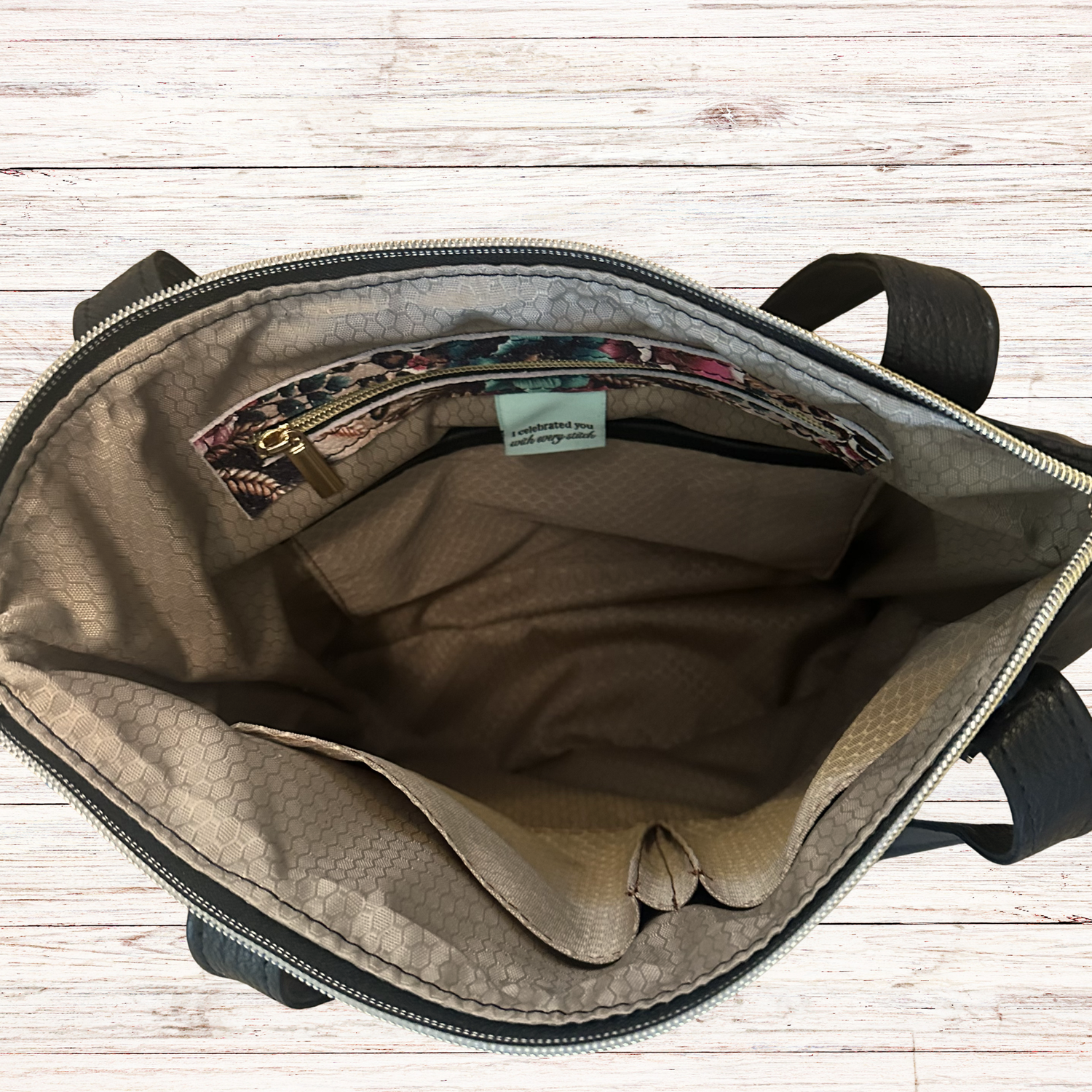 An open bag shown from above, revealing a beige and brown patterned interior with a zippered pocket and a small light blue label. The bag has black handles and is photographed on a light wood plank surface.