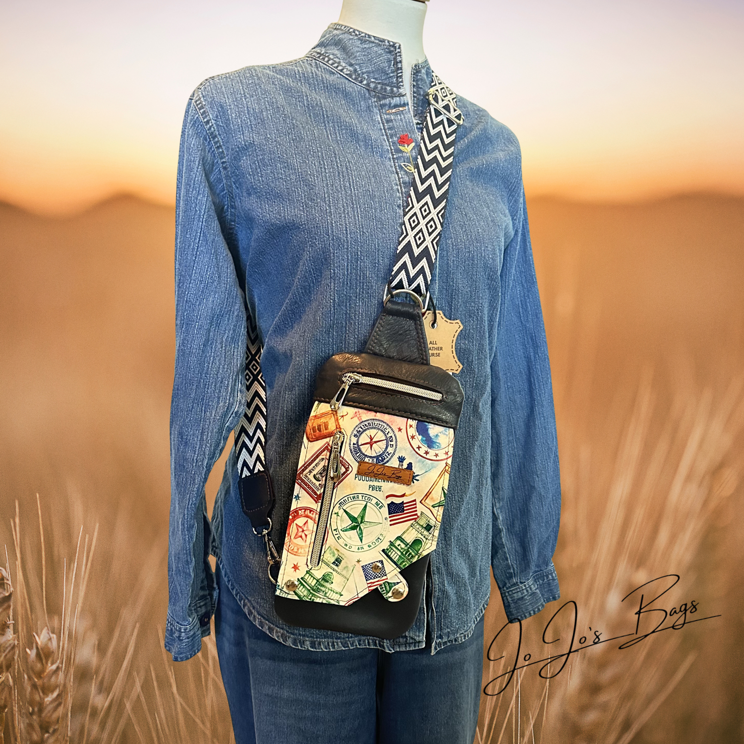 A mannequin wearing a blue denim shirt and jeans stands in a blurred wheat field at sunset, carrying a small crossbody bag with a colorful travel-postcard print and black trim. The brand name Jo Jo's Bags appears in script on the lower right.