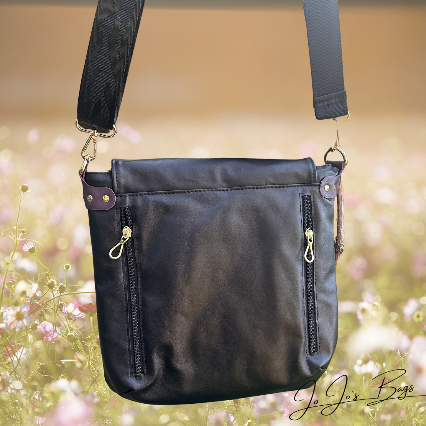 Black leather shoulder bag with a wide adjustable strap, two vertical zippered pockets, and gold-tone hardware, shown against a blurred outdoor floral background. A script logo reading 'JoJo's Bags' appears in the lower right corner.