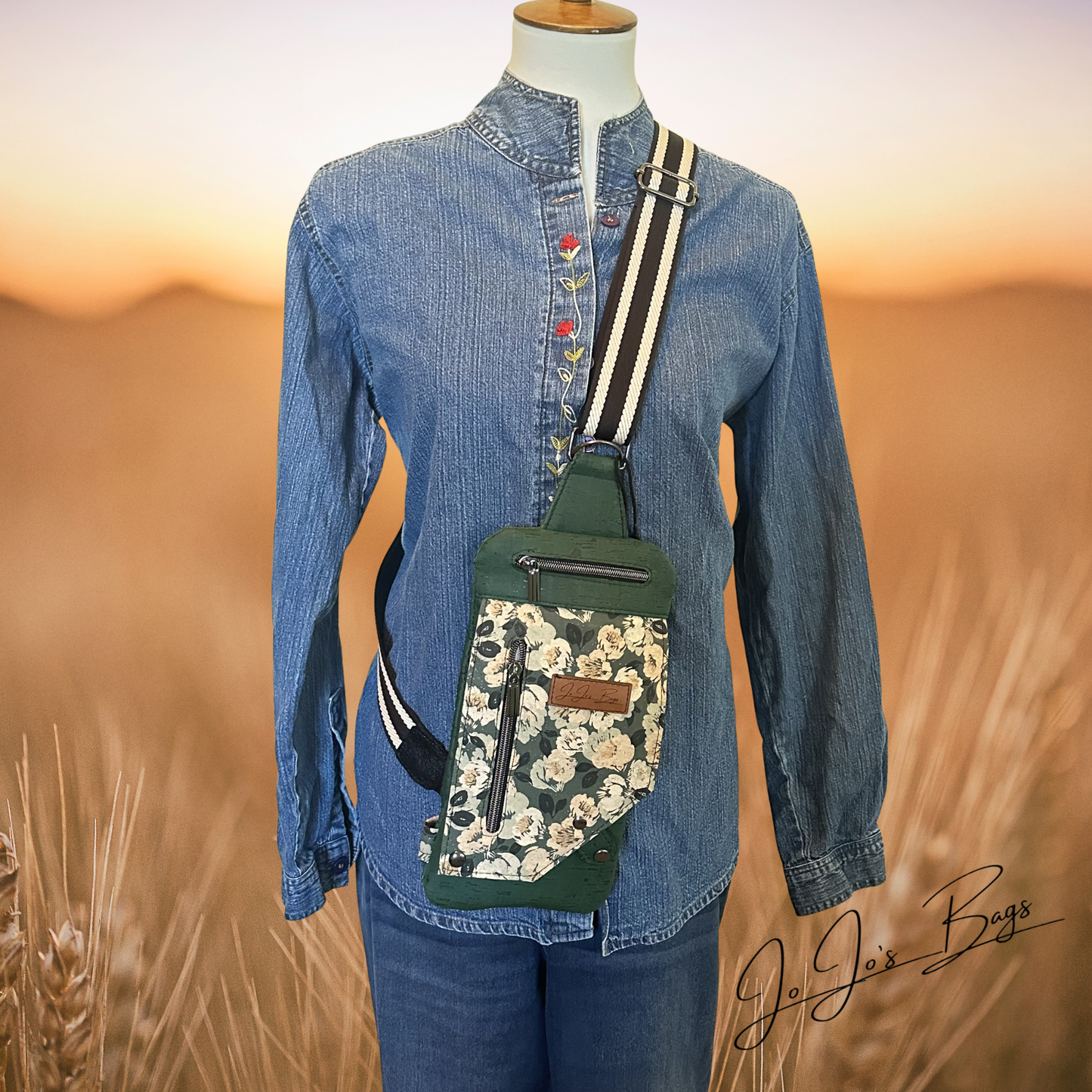 A mannequin wearing a blue denim shirt and jeans displays a green crossbody sling bag with a floral patterned front pocket and a striped strap, set against a blurred wheat field at sunset.