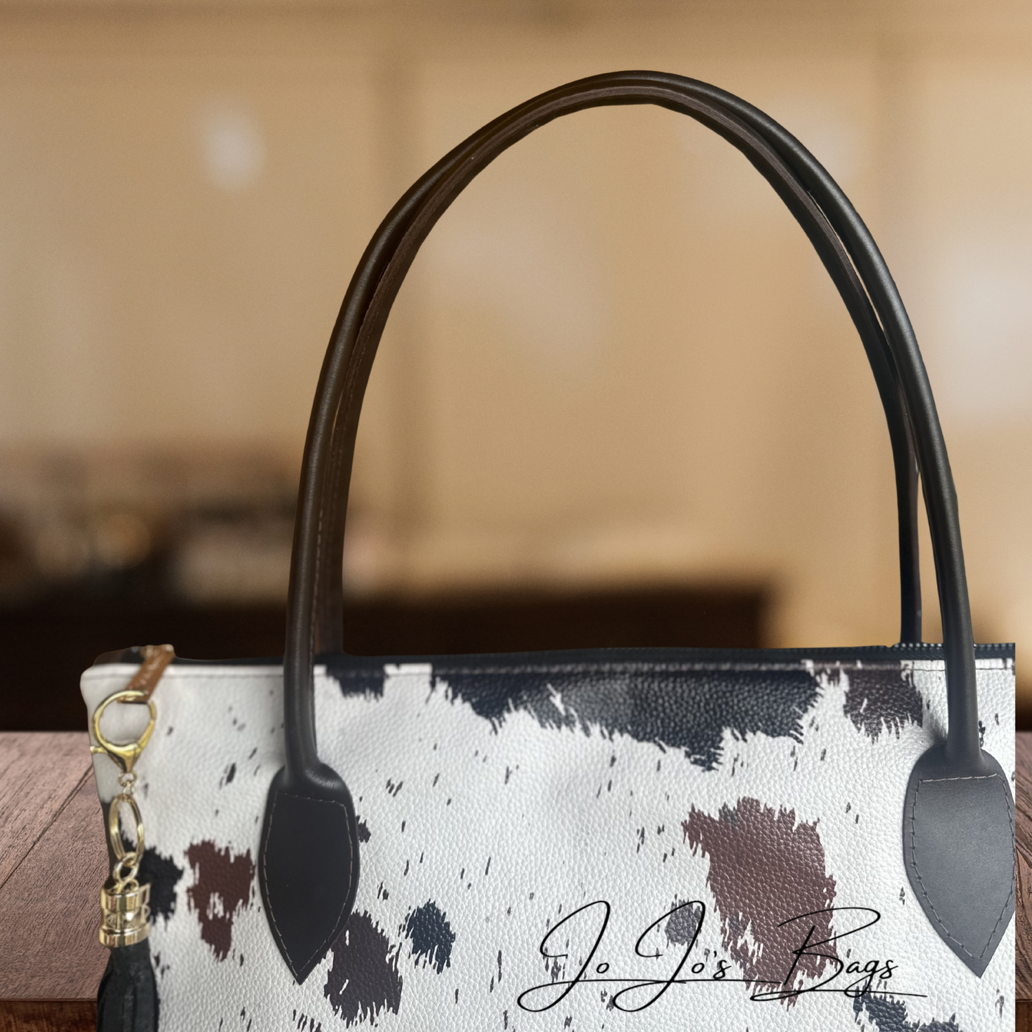 A close-up of a handbag with a white cow-print pattern, black and brown spots, and dark brown leather handles. A gold key ring and black tassel charm are attached on the left side.