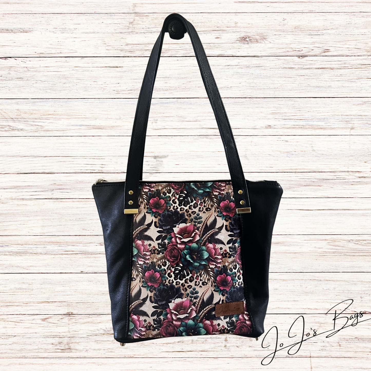 A black leather shoulder bag with a floral and leopard print front panel, displayed against a light wood plank background.