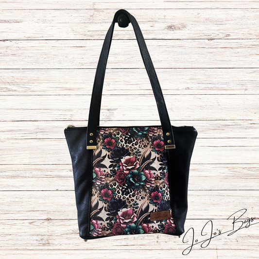 A black leather shoulder bag with a floral and leopard print front panel, displayed against a light wood plank background.