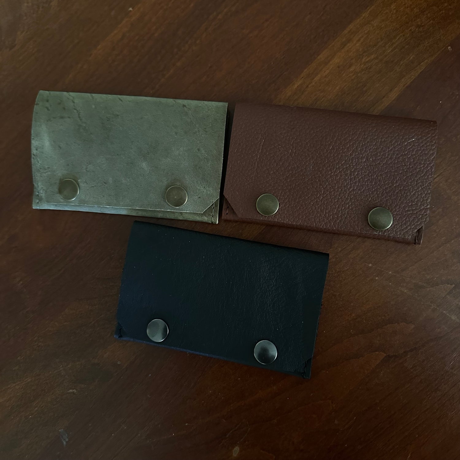 Three leather card holders in black, brown, and gray on a wooden surface.