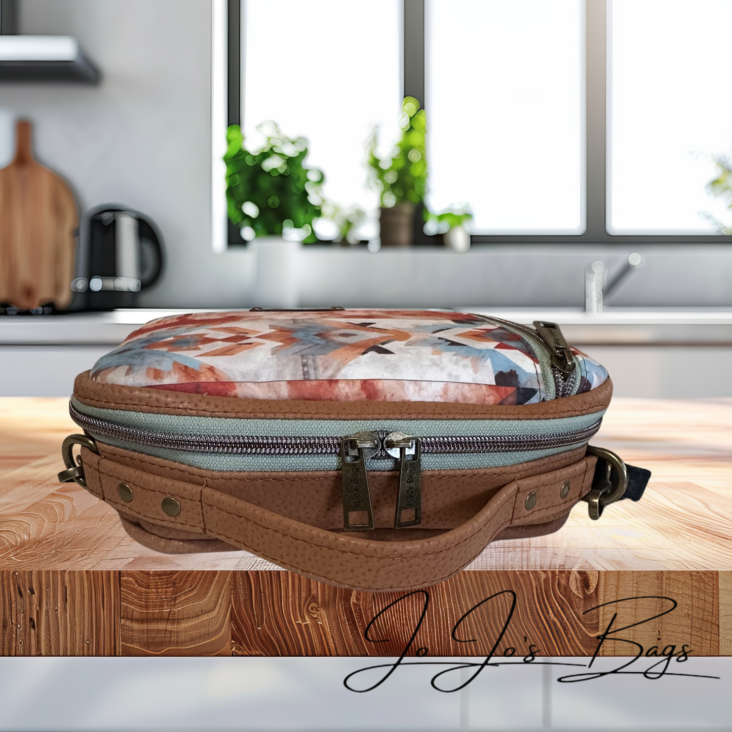 A brown leather crossbody bag with a colorful floral-patterned zippered pouch, resting on a wooden table in a blurred kitchen setting. The brand name 'JJ's Bags' is watermarked in the bottom right corner.