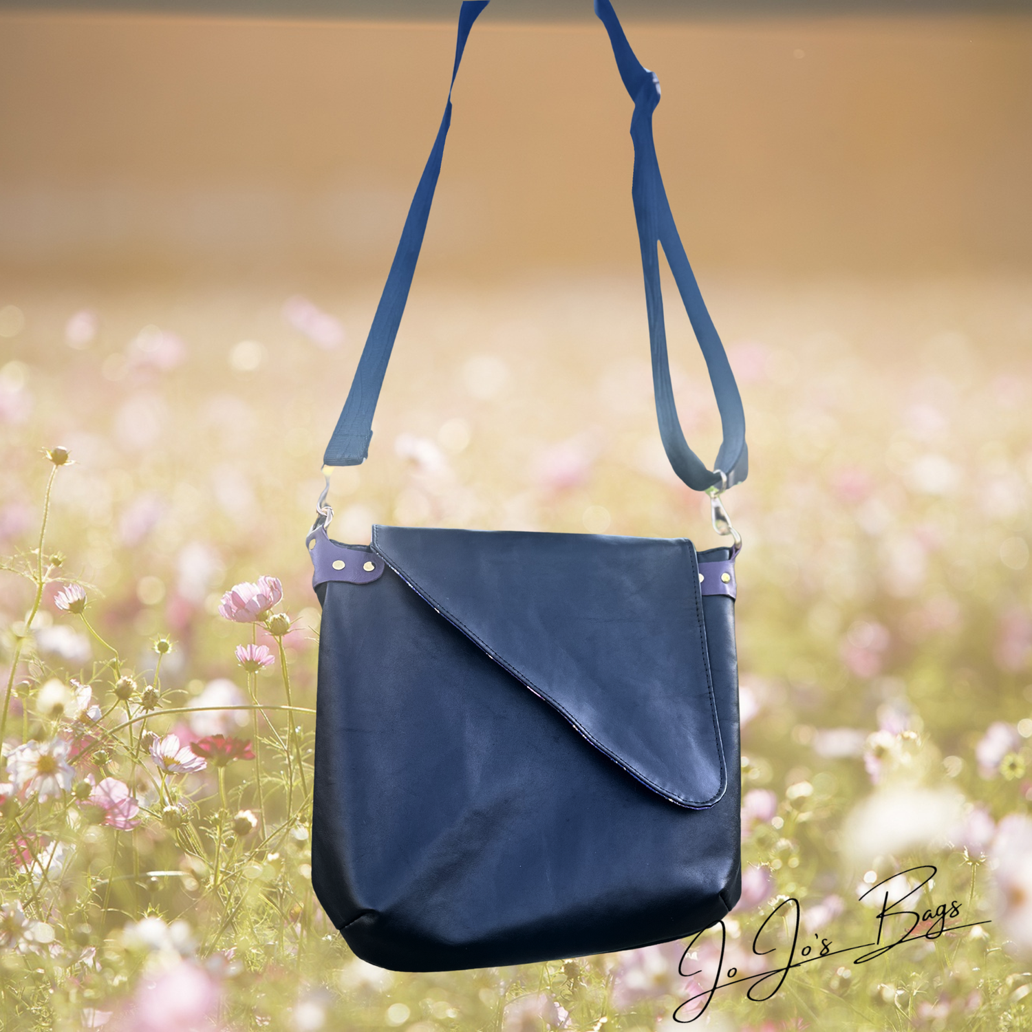 A black leather shoulder bag with a wide adjustable strap, displayed against a soft-focus background of a blooming flower field. The brand name 'Jo Jo's Bags' is visible in the bottom right corner.