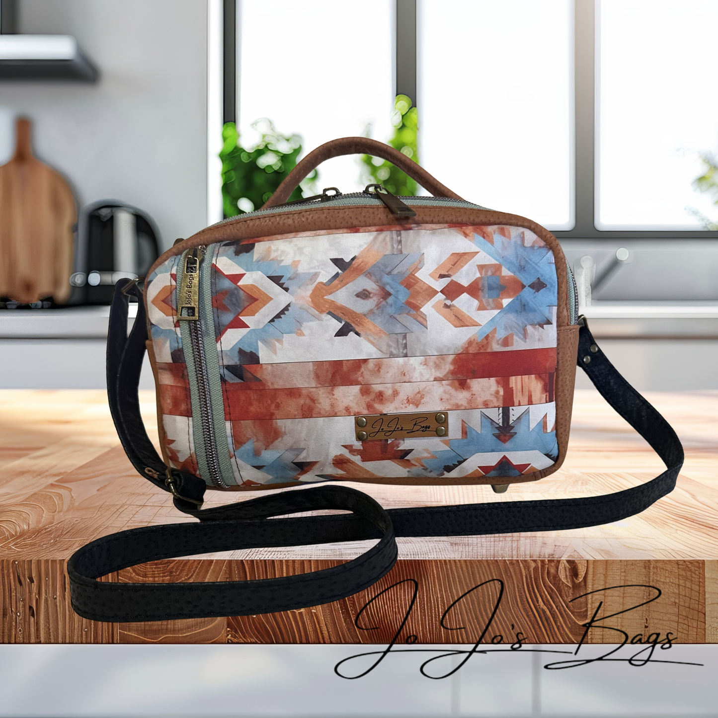 A patterned crossbody bag with a geometric Southwestern design in blue, orange, red, and white, featuring brown leather trim and a black shoulder strap, displayed on a wooden surface with a blurred kitchen background.