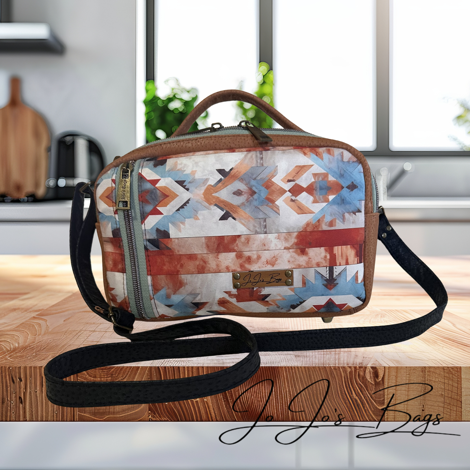 A patterned crossbody bag with a geometric Southwestern design in blue, orange, red, and white, featuring brown leather trim and a black shoulder strap, displayed on a wooden surface with a blurred kitchen background.