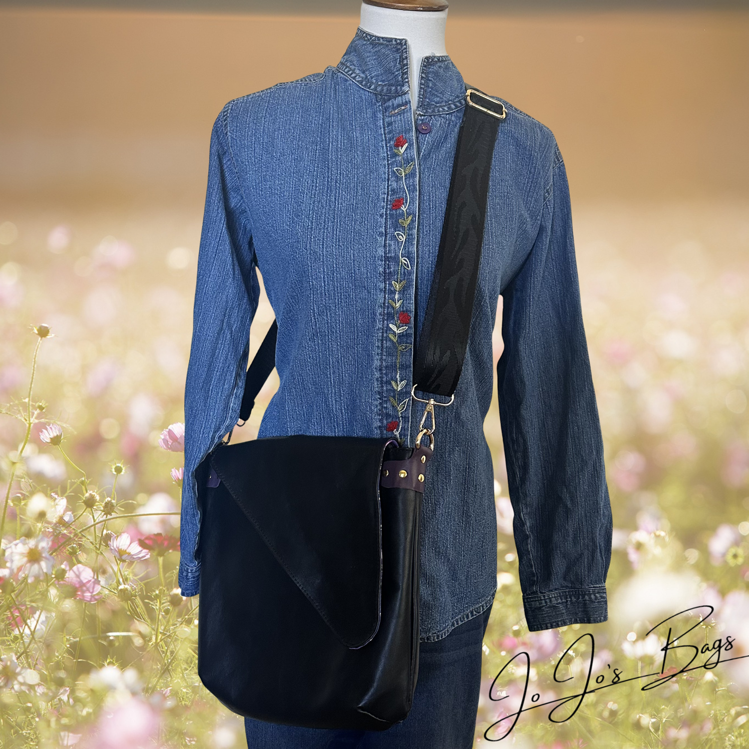 A black leather shoulder bag with a wide adjustable strap, displayed on a mannequin wearing a blue denim shirt against a blurred pink and yellow floral background. A 'Jo Jo's Bags' logo is visible in the bottom right corner.