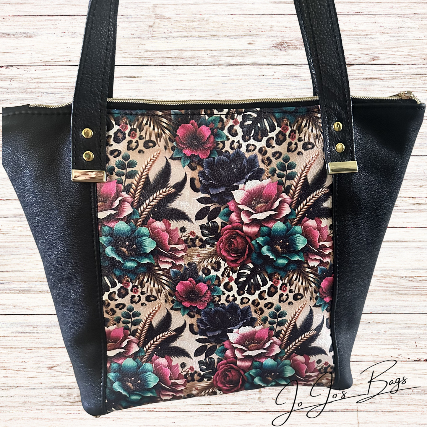 A black leather bag with black faux-leather sides and handles, featuring a central floral and leopard-print panel in pink, teal, black, and beige, photographed on a light wood background.