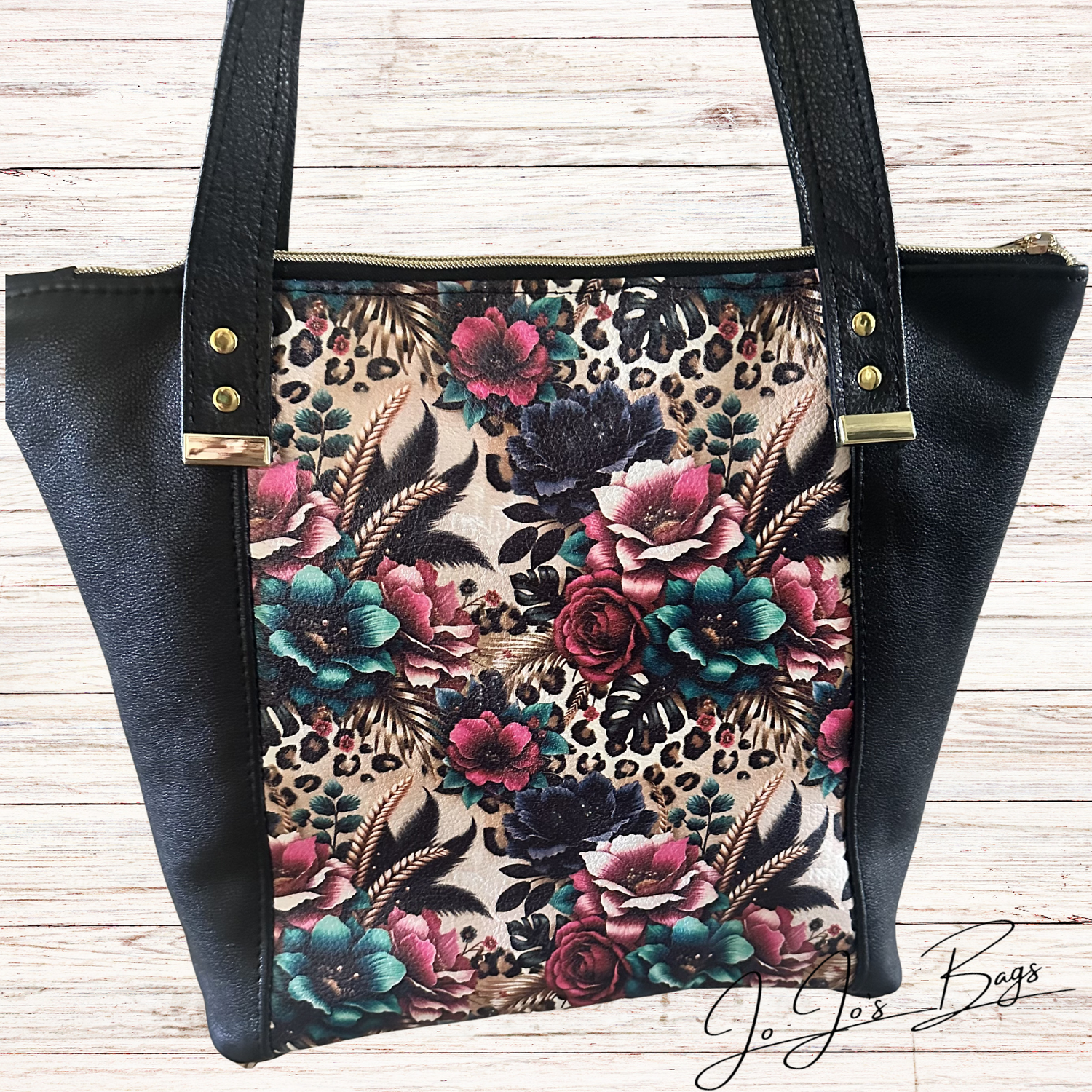 A black leather bag with black faux-leather sides and handles, featuring a central floral and leopard-print panel in pink, teal, black, and beige, photographed on a light wood background.