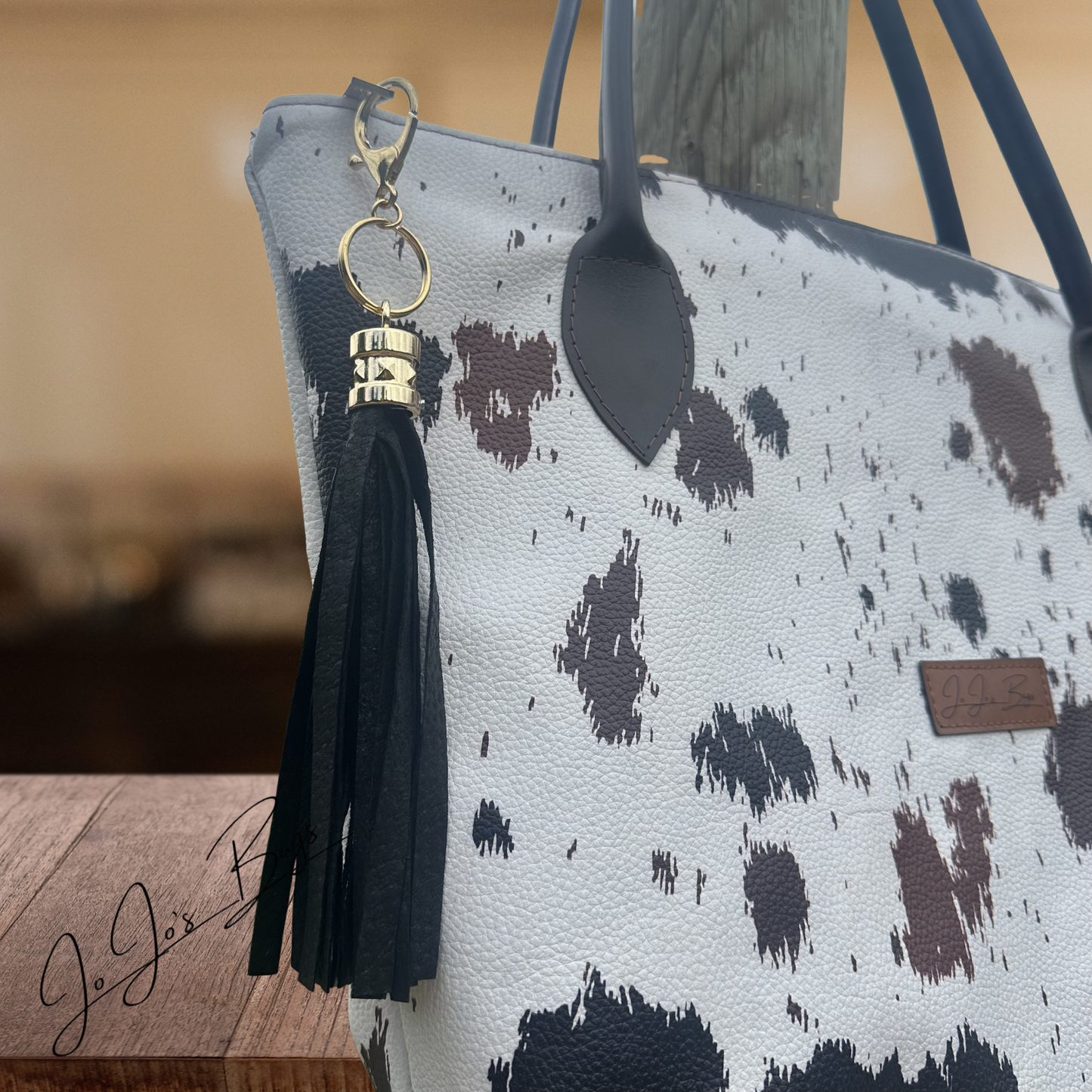 A close-up of a tote bag with a white pebbled texture, black and dark brown cow-print spots, dark gray handles, and a black tassel keychain with gold-tone hardware hanging from the side.