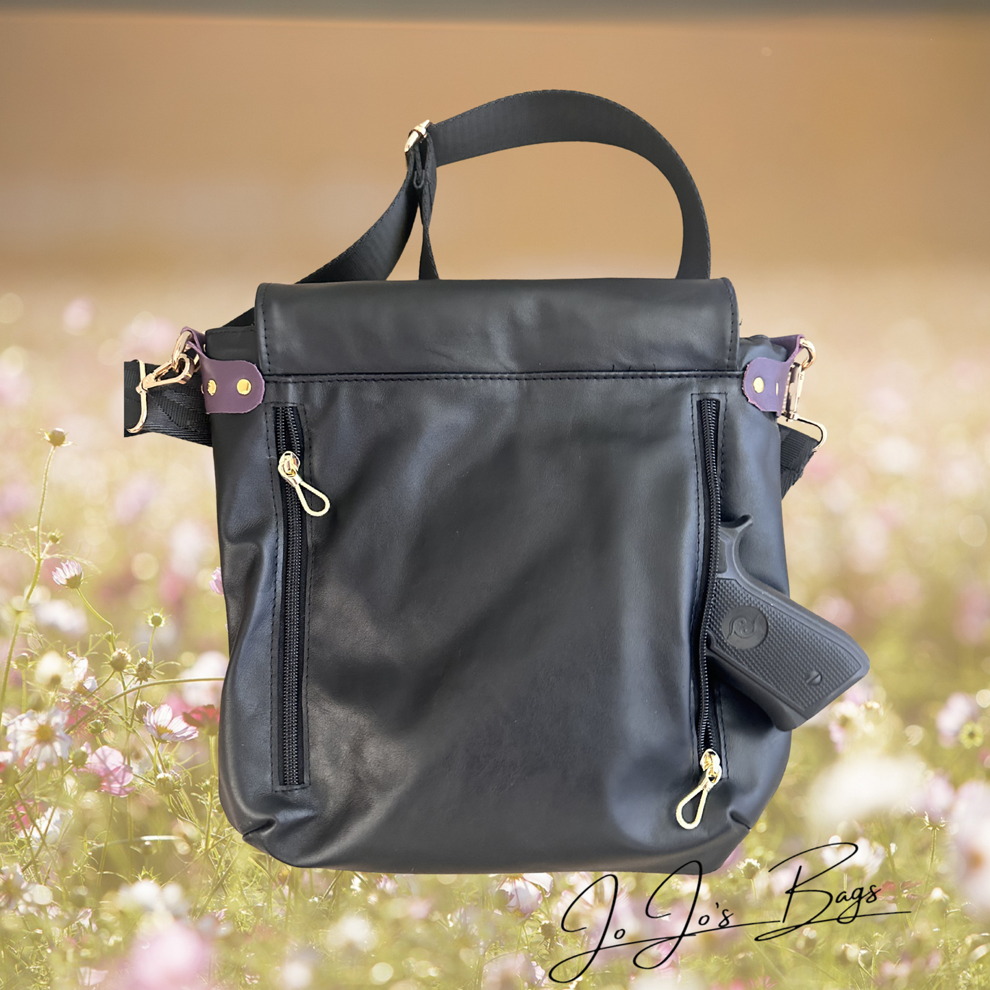 A black leather shoulder bag with a top handle, gold-tone hardware, and a handgun holstered on the side, set against a blurred field of pink and white flowers.