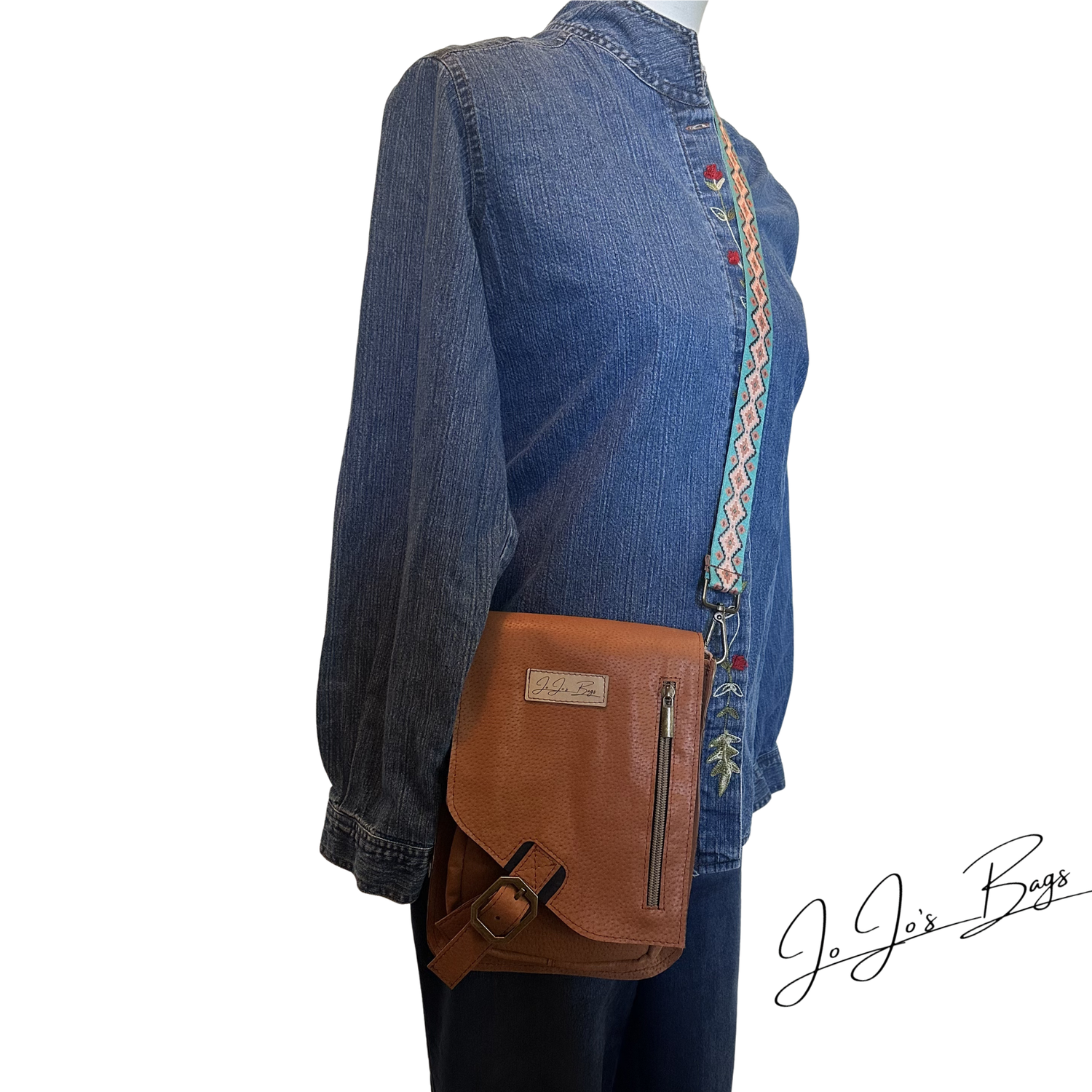 A brown leather crossbody bag with a front flap, buckle strap, and vertical zipper, shown worn across the torso of a mannequin dressed in a blue denim shirt. The bag has a small metal brand plaque and a patterned strap detail.