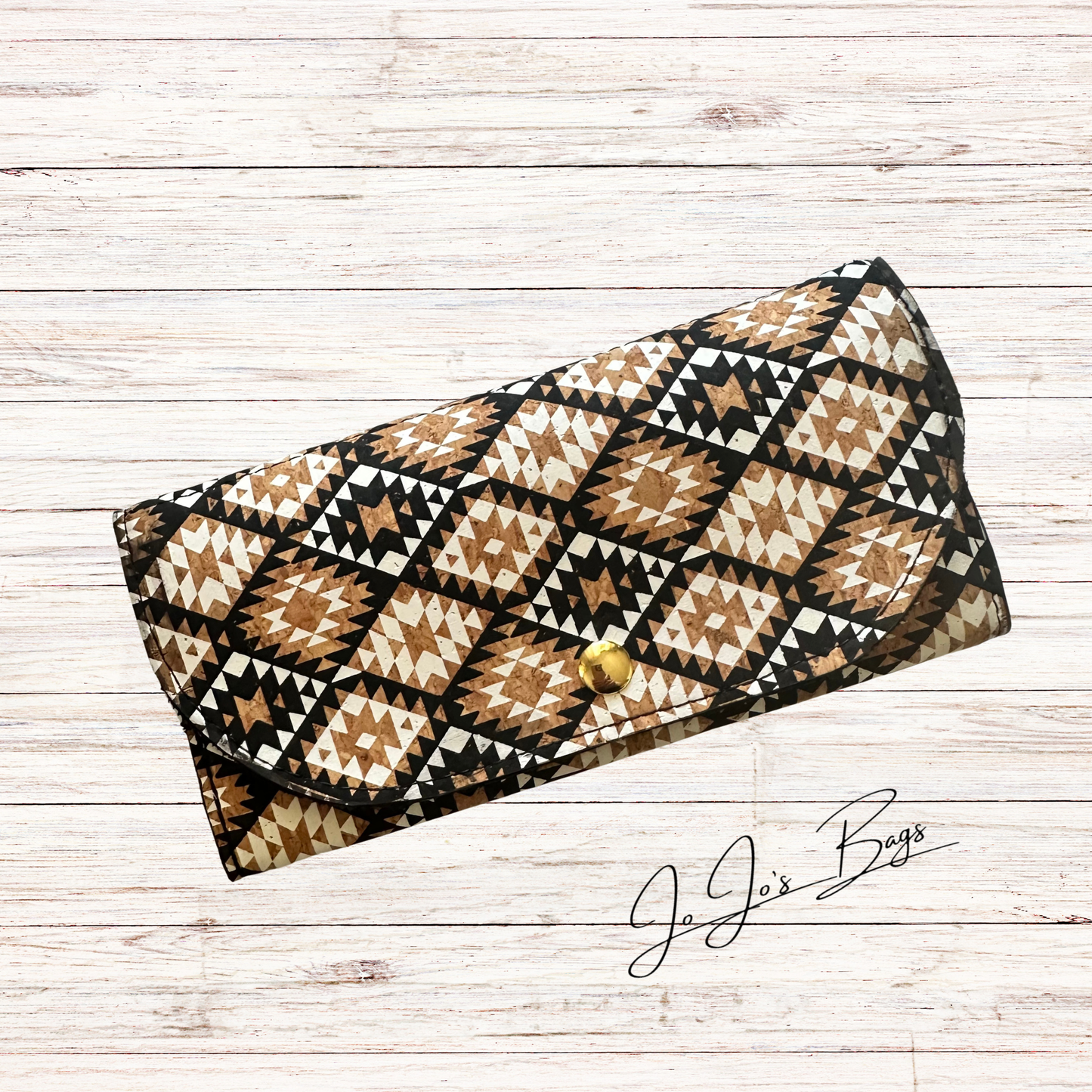 A long cork wallet with a Southwestern geometric pattern in brown, beige, black, and white, featuring a small gold button closure. The bag is displayed on a light whitewashed wood background with a 'Jo Jo's Bags' logo in the bottom right corner.