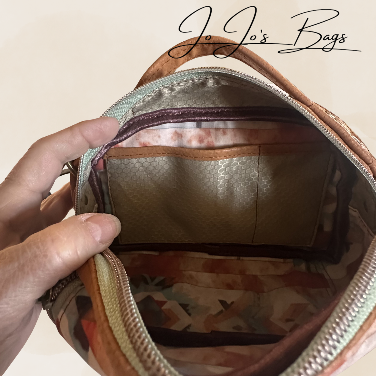 An open bag or pouch shown from the inside, with a hand holding the zipper area. The bag has a tan leather-like exterior, a patterned interior in beige, brown, and muted orange tones, and a visible JoJo's Bags logo at the top.