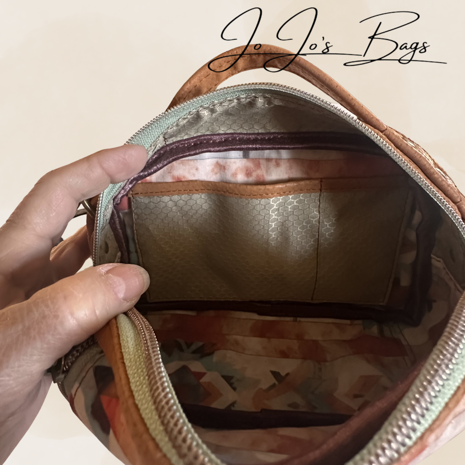 An open bag or pouch shown from the inside, with a hand holding the zipper area. The bag has a tan leather-like exterior, a patterned interior in beige, brown, and muted orange tones, and a visible JoJo's Bags logo at the top.