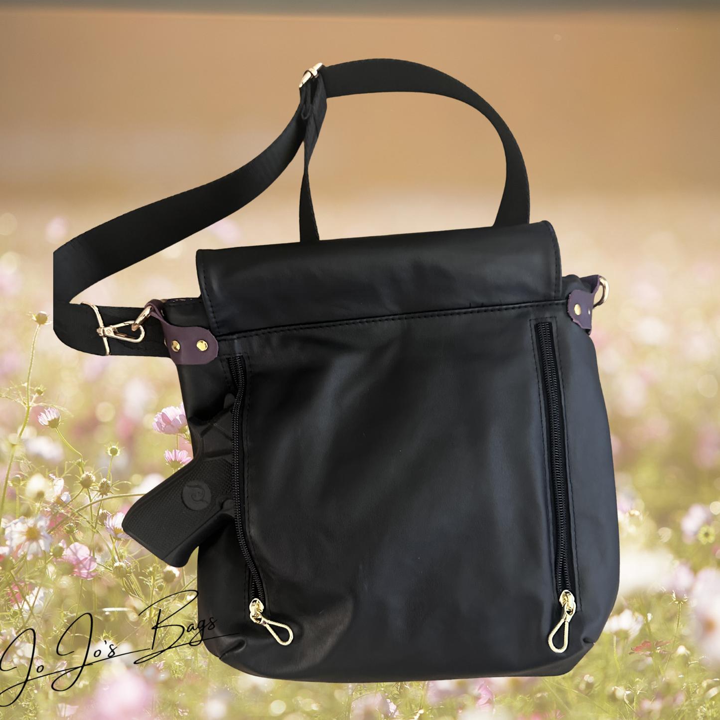 A black leather bag with a top handle, adjustable shoulder strap, and back zipper pocket, shown against a blurred outdoor floral background. A partial view of a pistol is seen. 