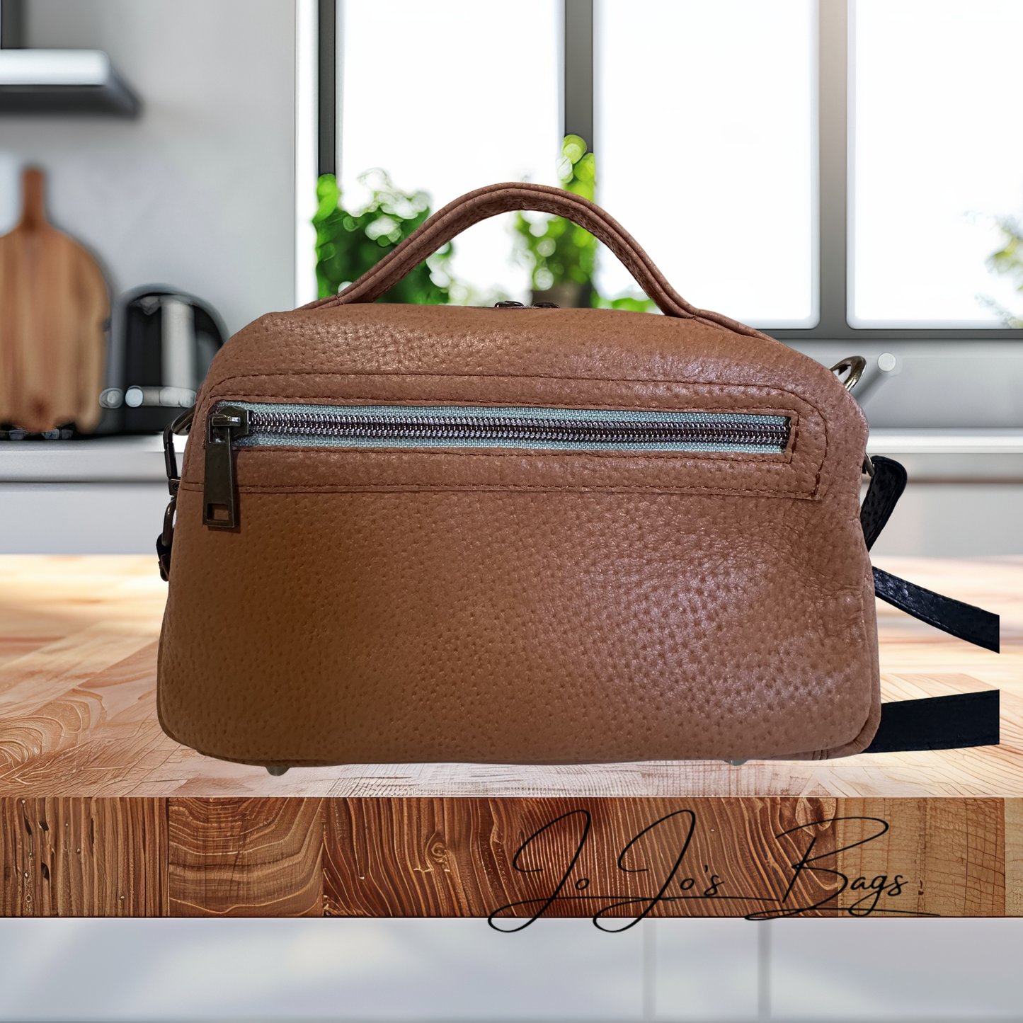 A brown pebbled leather handbag with a top handle and a front zippered pocket, displayed on a wooden surface with a blurred kitchen background. A 'Jo Jo's Bags' watermark is visible at the bottom right.