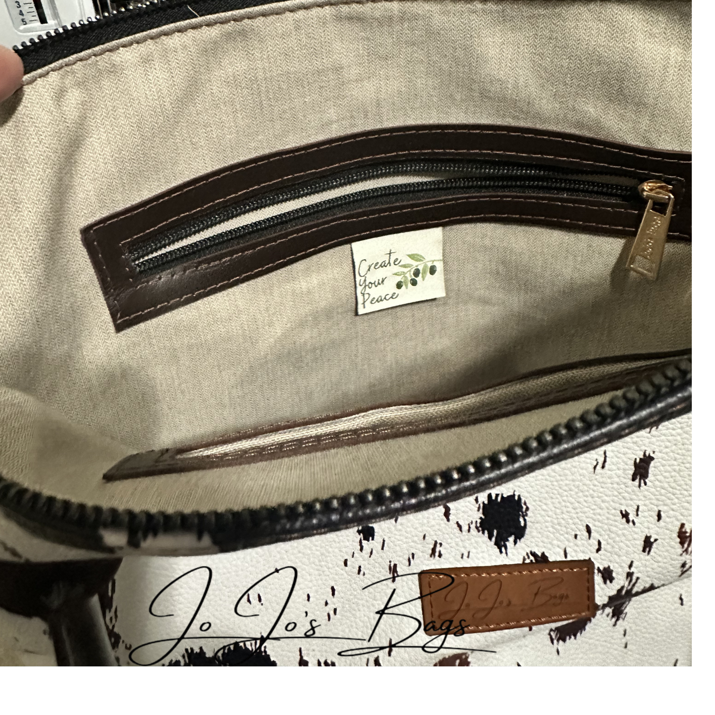 Close-up of the interior of a handbag showing a beige fabric pocket with dark brown trim and a small sewn label reading 'Create Your Passion.' A partial exterior view at the bottom shows a white and brown print leather surface with black splatter-like print and a brown leather patch.