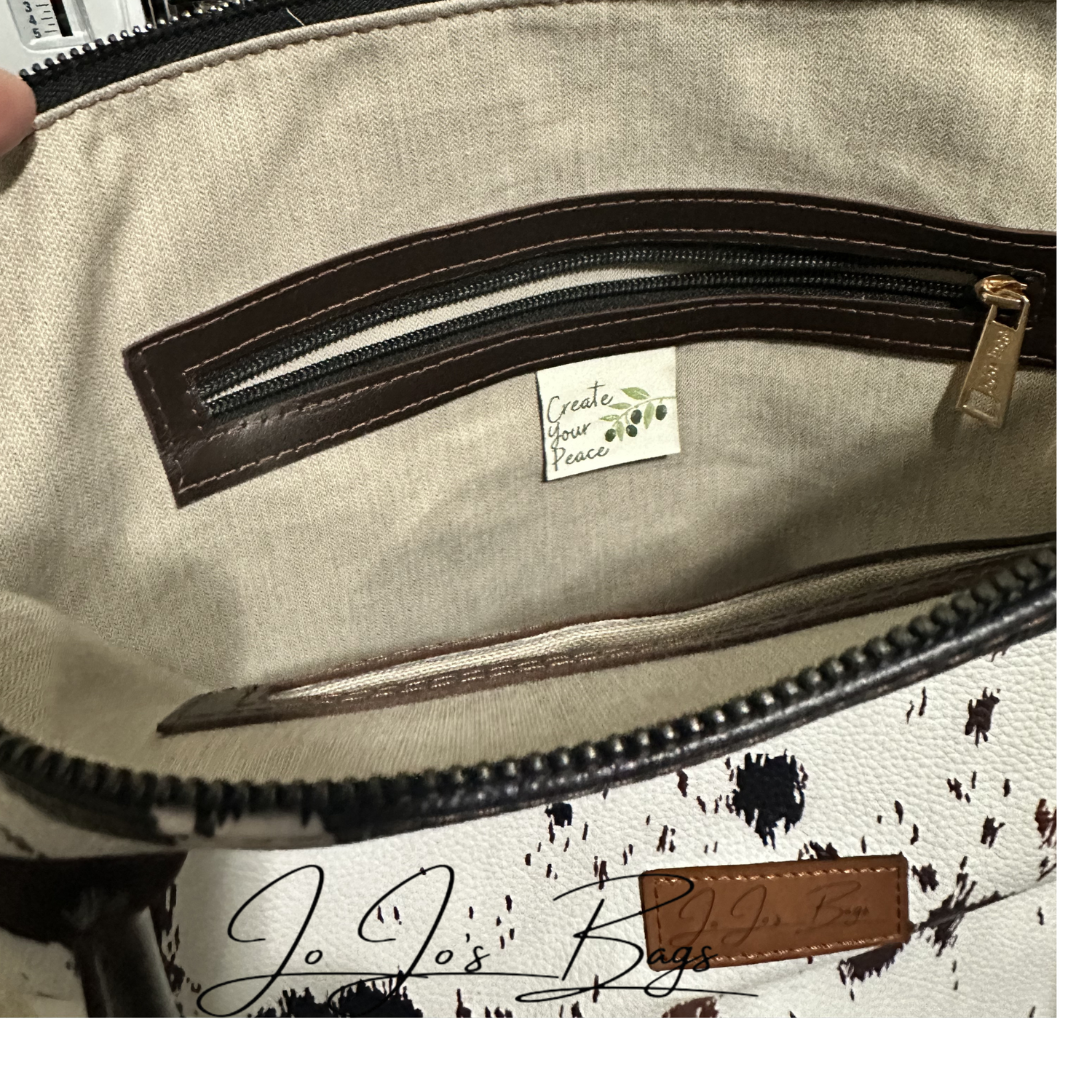 Close-up of the interior of a handbag showing a beige fabric pocket with dark brown trim and a small sewn label reading 'Create Your Passion.' A partial exterior view at the bottom shows a white and brown print leather surface with black splatter-like print and a brown leather patch.