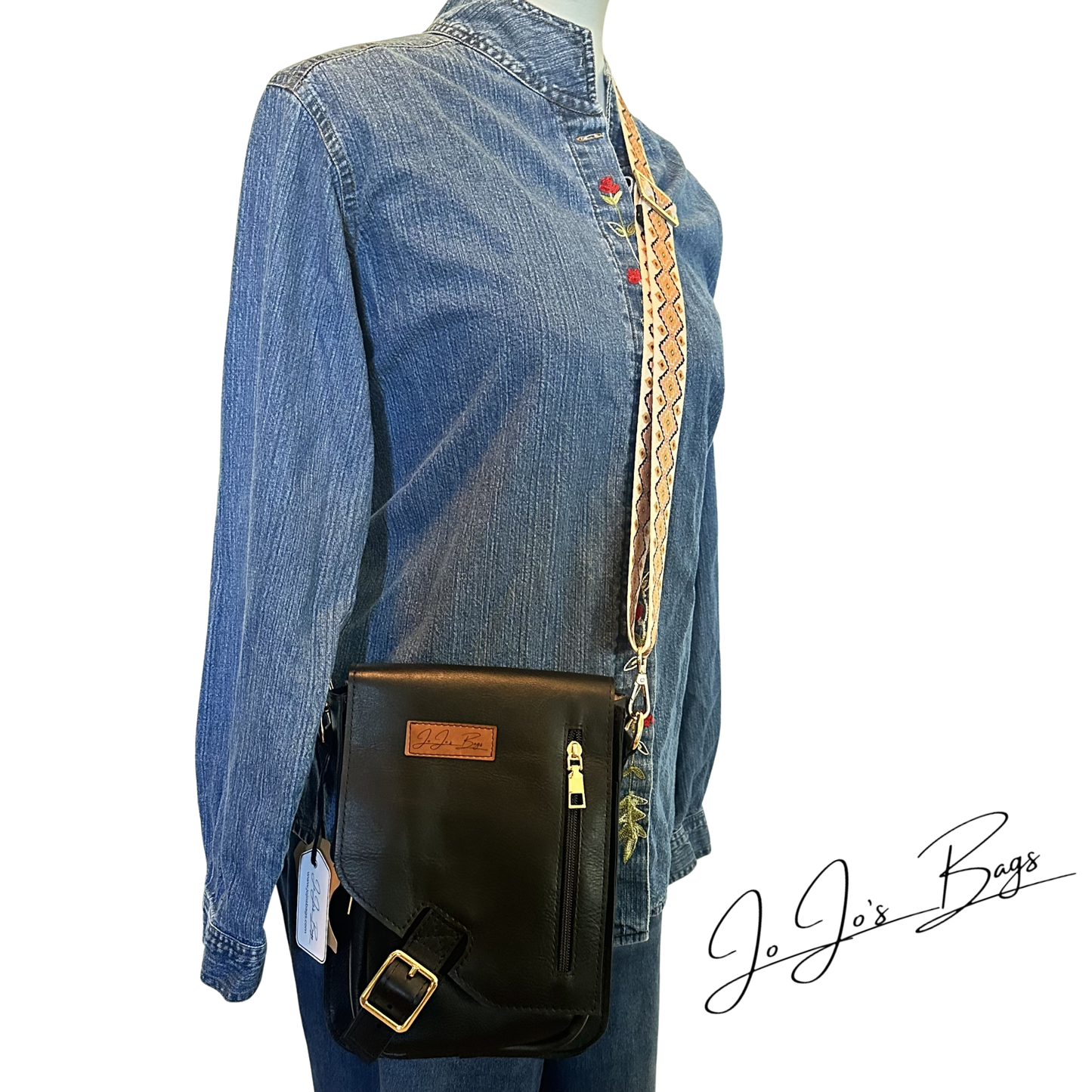 A black leather crossbody bag with a gold-tone buckle and a shoulder strap, shown worn across a blue denim shirt on a mannequin. The brand name 'JoJo's Bags' is written in script on the bottom right.