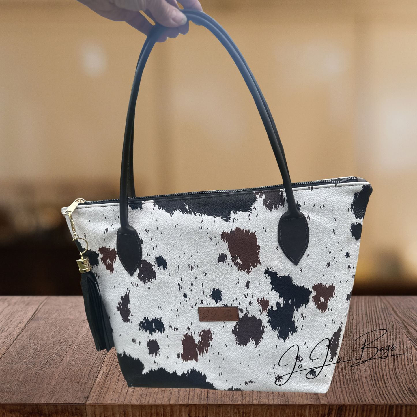 A cow-print handbag with black leather handles and a black tassel charm, held by a hand over a wooden table.