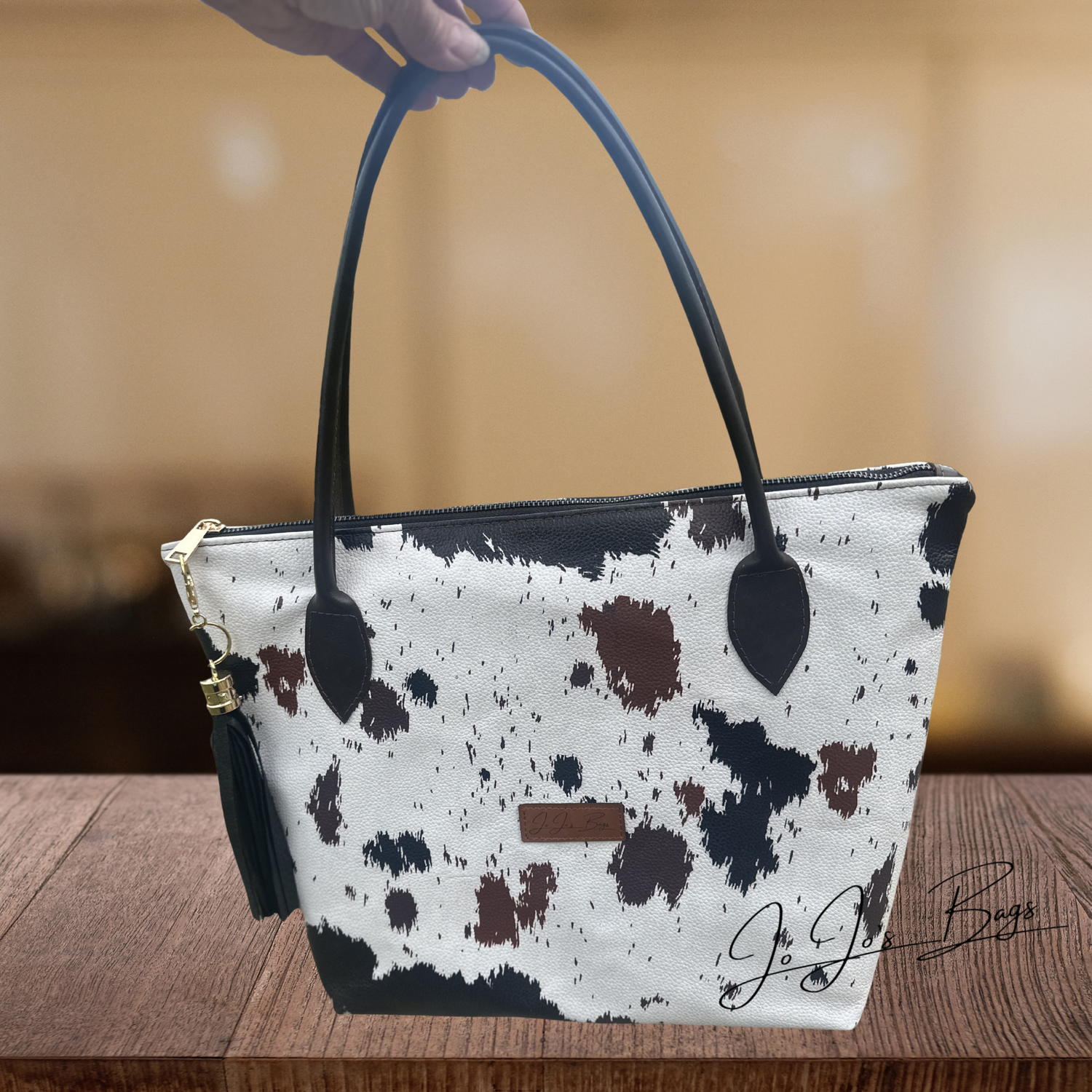 A cow-print handbag with black leather handles and a black tassel charm, held by a hand over a wooden table.