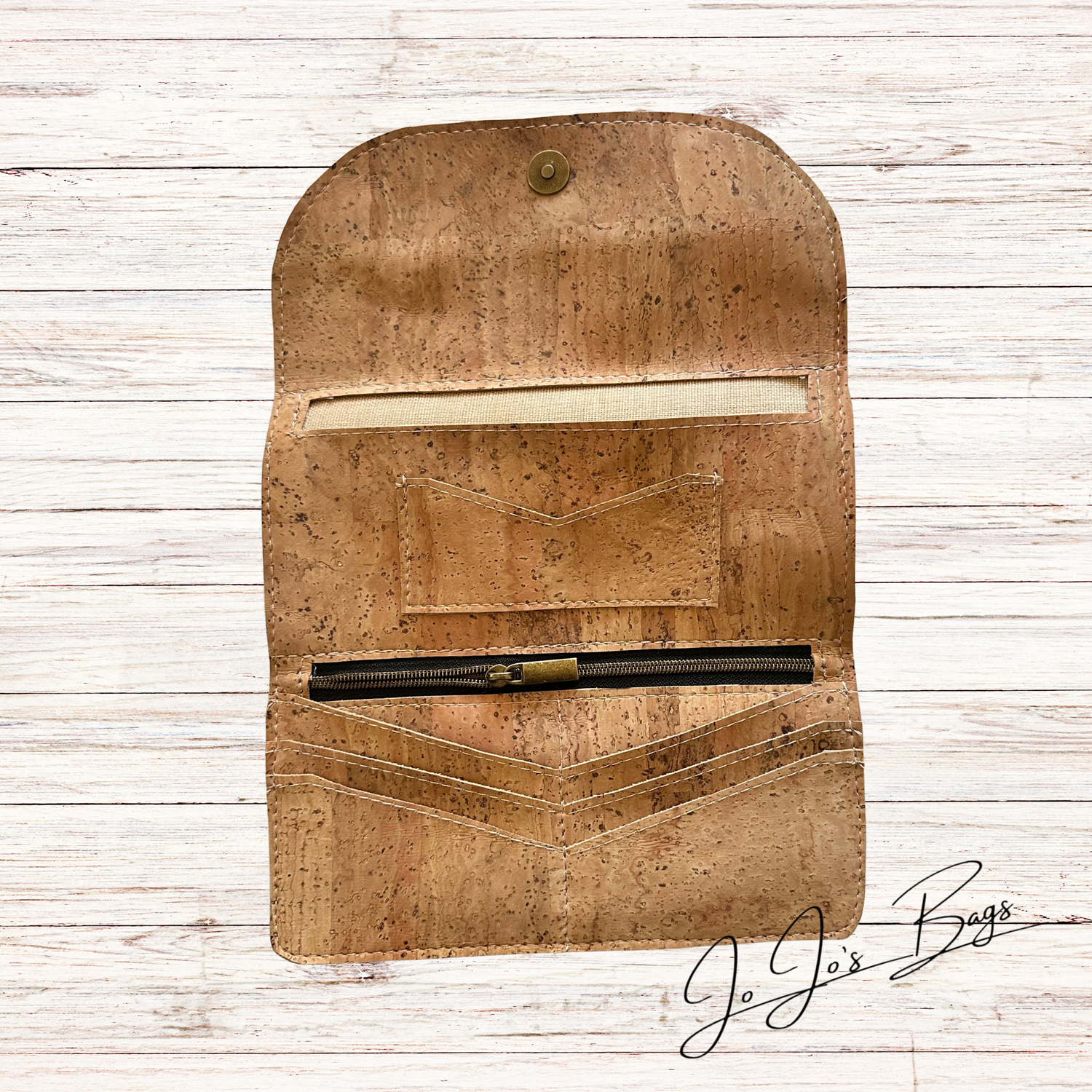 A cork wallet with a snap button closure, shown open on a white wood plank background. The wallet features a zippered pocket holding a black pen, card slots, and a cream-colored interior lining.